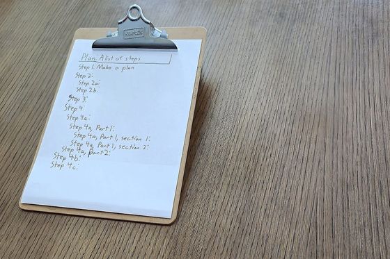 Klemmbrett mit handschriftlicher Schrittliste „Plan: A list of steps“ auf einem Holztisch