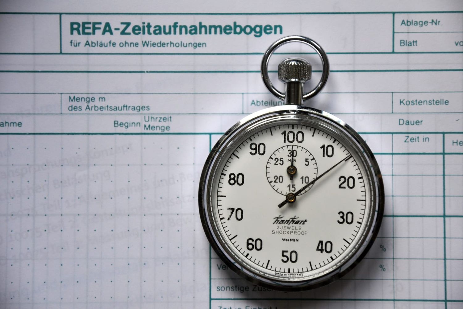 Mechanische Stoppuhr auf einem Zeitaufnahmebogen mit Spalten für Dauer und Kostenstelle